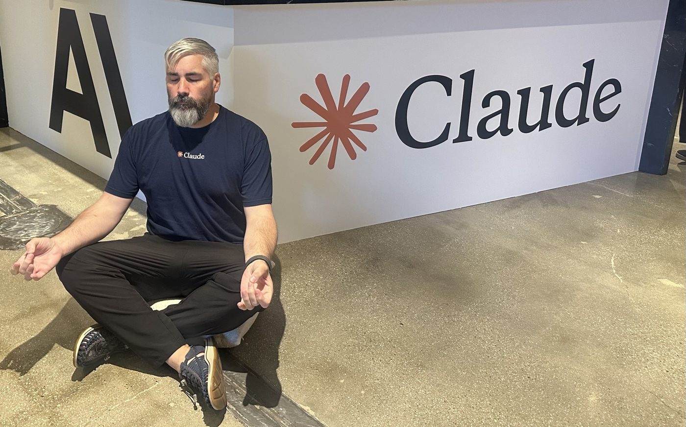 Jeremy Curtis sitting cross-legged in a meditation pose in front of a large Claude sign